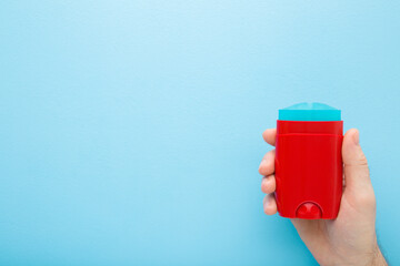 Young adult man hand holding and showing red plastic stick of armpit deodorant on light blue table...