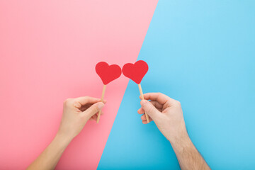 Young adult woman and man hands holding red paper heart shapes on wooden sticks on light pink blue...