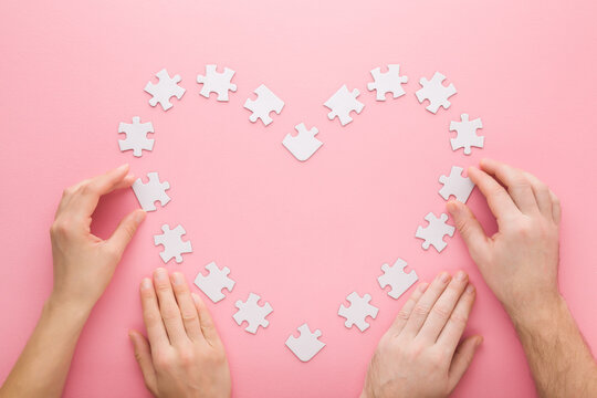 Young adult woman and man hands assembling white heart shape puzzle pieces on pink table background. Pastel color. Closeup. Love and relationship concept. Couple spending time together. Top down view.