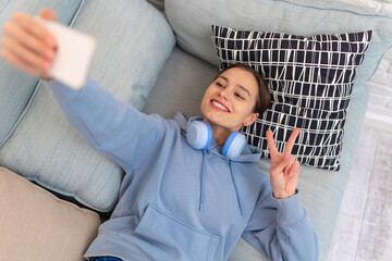 Woman with headphones making selfie on couch showing v sign in home interior.
