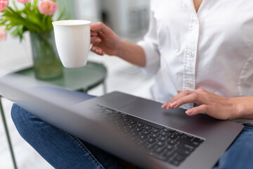 Woman holding cup with coffee working on her laptop remotely from home office.