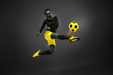 Competitive young woman, soccer playing in motion, hitting ball with leg, playing over black background. Yellow elements. Monochrome. Concept of competition, tournament, match, game. Creative design.