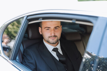 a handsome groom sits with a wedding bouquet inside a white car and looks out the window. A happy moment on the wedding day.