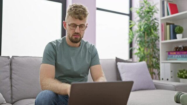 A young man with a beard and glasses feeling stomach pain while using a laptop on a sofa indoors.
