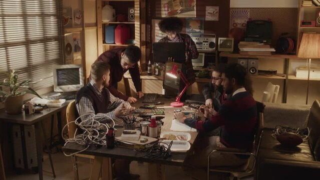 Lowkey shot of team of ethnically diverse male engineers working with circuit boards soldering and fixing them, sitting at desk with various wires in small homemade retro office with dimmed light