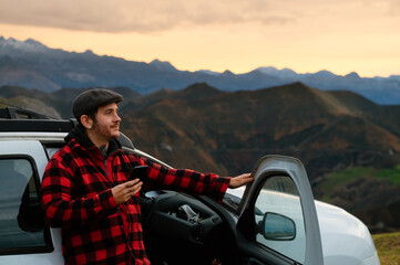 young man with beard and beret leaning on the door of his off-road car while using his smartphone in the mountains. He is using a map application on his phone to guide him on his trip. 