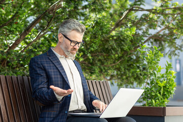Mature male financier in a smart suit using a laptop outside an office building, exuding professionalism and experience.