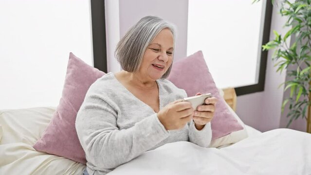 A smiling senior woman uses a smartphone while relaxing in her bedroom, depicting technology use by the elderly at home.