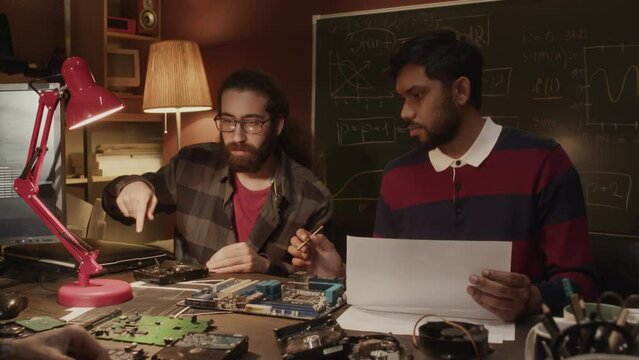 Waist up of ethnically diverse team of computer engineers having discussion while repairing circuit boards at desk in dark old-fashioned garage workshop