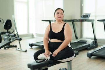 Overweight Asian woman exercise in gym,  Smiling young woman clothes sitting on workout bench, enjoying a break. Plus-size gym-goer happily using phone after exercise, radiating positivity