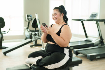 Fototapeta na wymiar Overweight Asian woman exercise in gym, Joyful in activewear using smartphone, resting on bench with treadmills in behind. Smiling plus-sized female casually seated on bench, itness machines behind