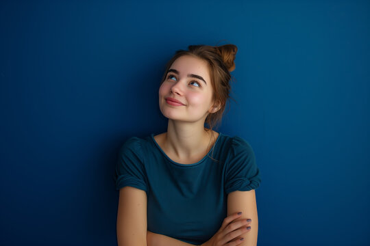 Young Woman Smiling and Looking Upwards. A young woman with a top bun hairstyle smiles while looking upwards, against a vivid blue background, exuding optimism.
