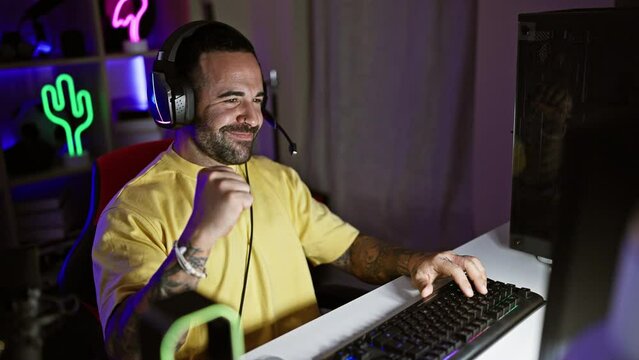 Handsome hispanic man with a beard wearing headphones in a neon-lit gaming room at night focusing on a computer screen.