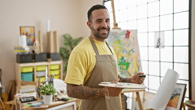 A confident hispanic man with a beard stands in a bright studio holding a paintbrush and palette, wearing a yellow shirt and apron.