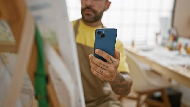 Hispanic tattooed man with beard using smartphone in art studio, portrait of focused adult male artist indoors.