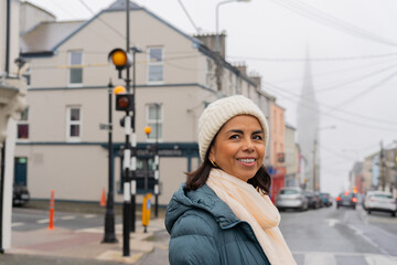 Obraz na płótnie Canvas Beautiful latin woman with a winter hat enjoying the charm of a picturesque town in Ireland with the cathedral in the background on her car trip through the country on a somewhat foggy day