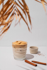 Dalgona coffee. Whipped instant iced coffee with milk in a glass with cinnamon sticks on gray background with palm leaf.