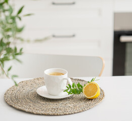 A cup of tea with lemon and a teapot and green branch on a white table against the background of a white kitchen.