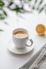 A cup of tea with lemon and green branch on a white table against the background of a kitchen window.