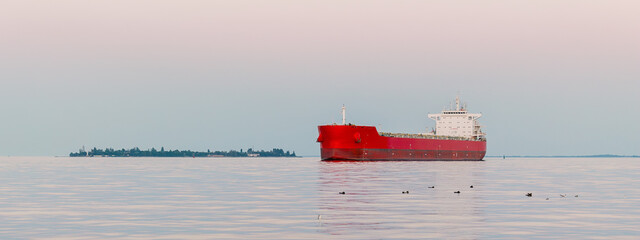 A red cargo ship is sailing on the sea next to the island.