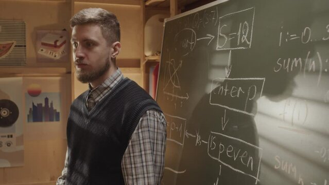 Medium shot of young Caucasian male startuper presenting his new program idea to team, pointing at scheme on chalkboard standing in garage office