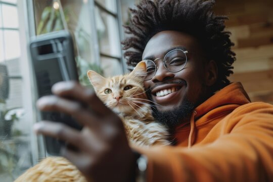 A young African American man with a beard and glasses takes a selfie with a ginger cat, both looking into the camera with a backdrop of indoor plants.