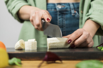 Woman cutting banana, preparing healthy vegan smoothie on a table