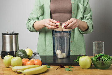 Woman holding banana pieces, preparing healthy vegan smoothie with spinach, apple and avocado in a blender on a table