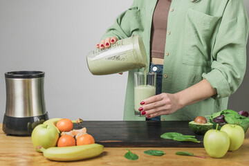 Unrecognizable woman in casual wear pouring fresh blended healthy green vegan smoothie into a...