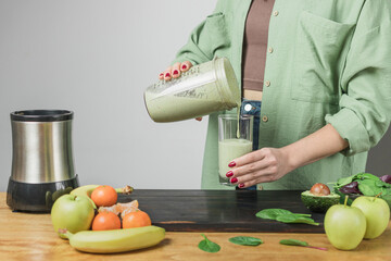 Unrecognizable woman in casual wear pouring fresh blended healthy green vegan smoothie into a...