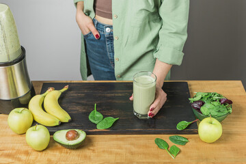 Unrecognizable woman in casual wear holding glass of fresh blended healthy green vegan smoothie. banana, spinach, apple and avocado on a table