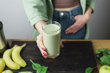 Unrecognizable woman in casual wear holding glass of fresh blended healthy green vegan smoothie. banana, spinach, apple and avocado on a table