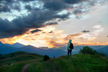 A girl tourist admires the sunset in the mountains. Silhouettes of mountains against the background of the sunset sky and a dirt road through a green meadow with wild flowers.