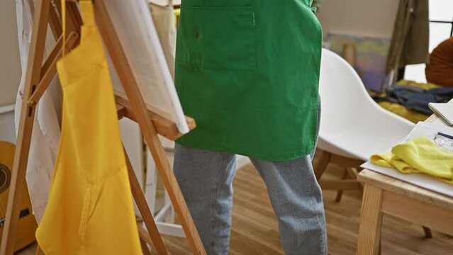 A focused young male artist in a green apron examines his palette in a bright art studio