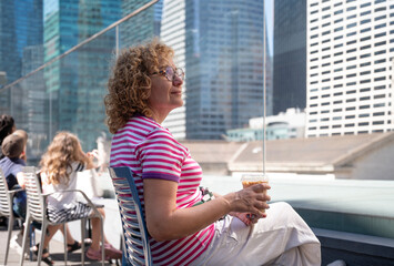 Adult woman enjoying a coffee on a sunny rooftop with skyscrapers in the background.