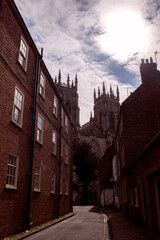 Fototapeta na wymiar Medieval York Majesty: Sunlit Alley, York Minster Peeking Behind Houses, Red Brick Charm, Tranquil Urban Scene with Blue Sky – A Timeless Glimpse of Historic England