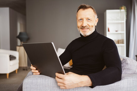 Smiling Bestager Man Sitting on Sofa with Laptop, Looking at Camera
