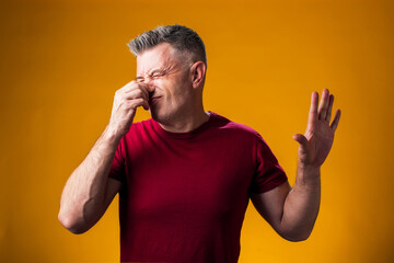 Man showing disgust and covering his nose with hand on yellow isolated background, guy refuses bad...