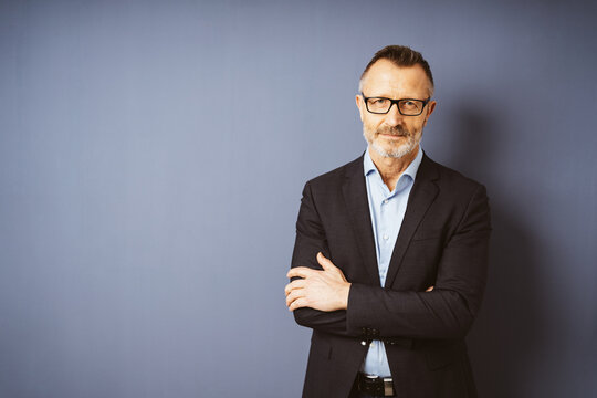 Serious Elderly Businessman with Eyeglasses Standing in Front of Blue Wall with Copy Space, Arms Crossed, Looking Sternly at the Camera
