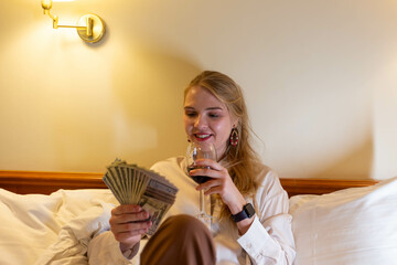 Smiling redhead woman counting money cash, use smartphone calculate domestic bills at vintage hotel room. Girl satisfied of income and saves money for planned vacation, gifts.