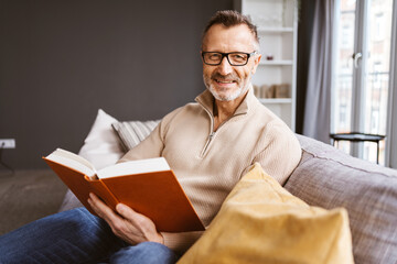 Middle-aged Man with Glasses Sitting Relaxingly on Sofa, Holding a Book, and Smiling at the Camera