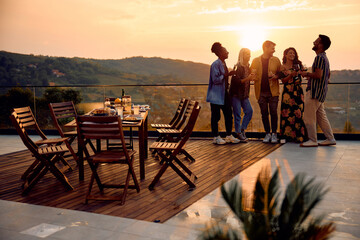 Cheerful friends have fun while drinking wine and talking on terrace at sunset.
