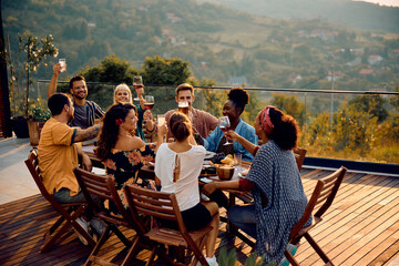 Multiracial group of happy friends toasting while gathering at dining table on terrace.