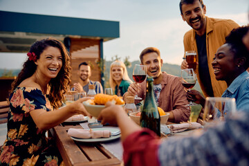 Cheerful woman and her friends enjoy in food and drinks while gathering at dining table on terrace.