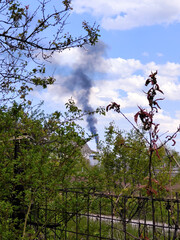 Behind the trees of the park, black smoke is coming from the plant's pipes against the background of the blue sky