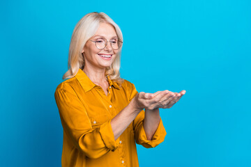 Photo portrait of lovely retired lady hold palms empty space dressed stylish orange smart casual garment isolated on blue color background