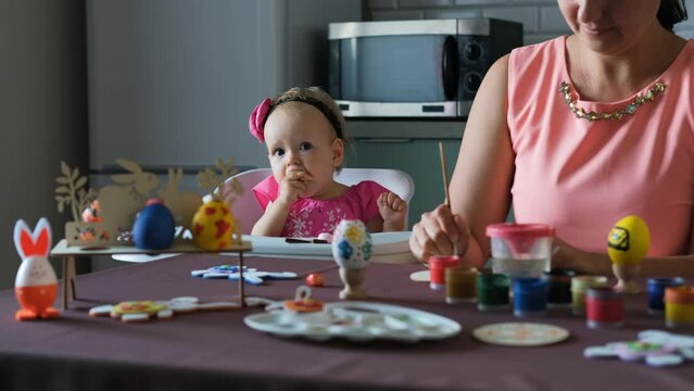 Adorable baby girl in pink dress eating cookie while her mother painting Easter egg at table in the kitchen at home