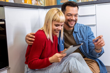 Happy couple sitting on the kitchen floor and ordering something online with their credit card