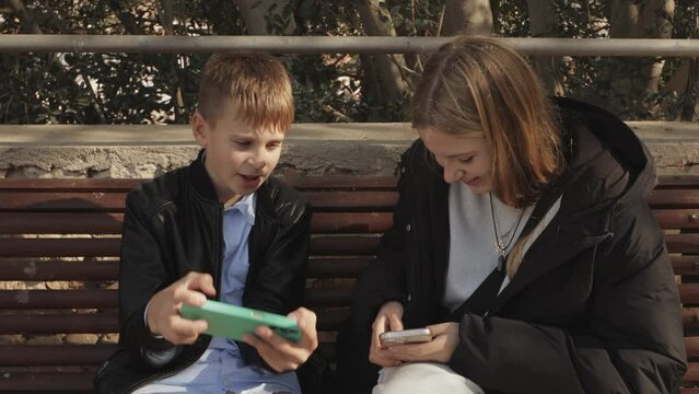 happy brother and sister sitting on a bench in the park holding phones in their hands, children 