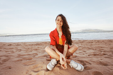 Fototapeta na wymiar Cheerful Woman Sitting on Sandy Beach, Enjoying Vacation and Smiling Wide in Trendy Fashion Outfit